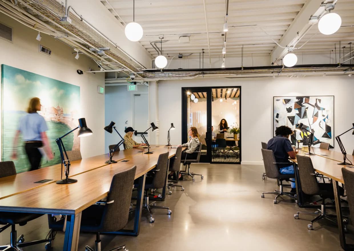A row of desks with task lamps under globe lights, with a glass meeting room at the back